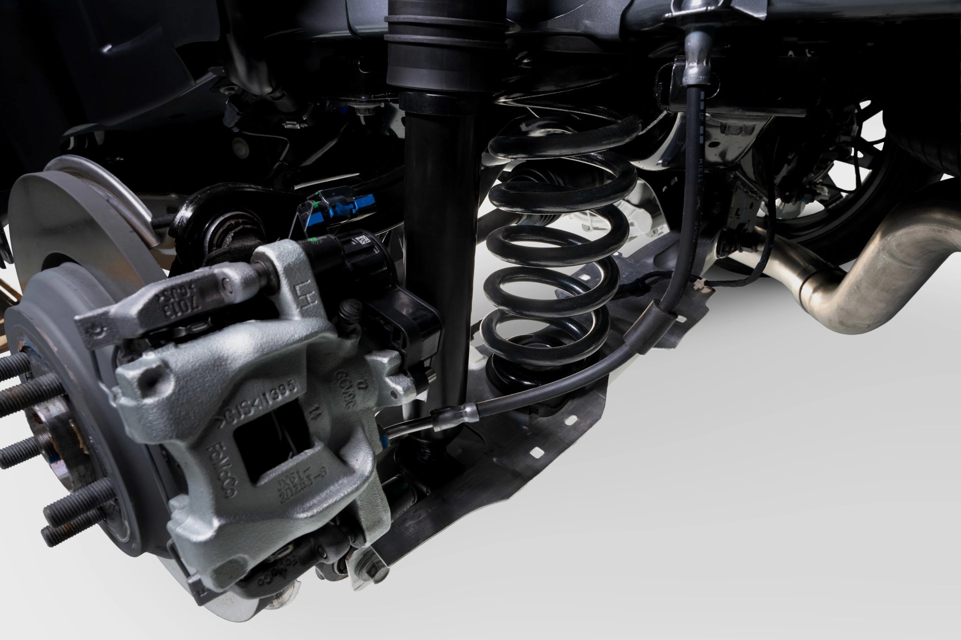 Close-up of a car suspension system showing a black coil spring, silver brake caliper, and exhaust pipe.