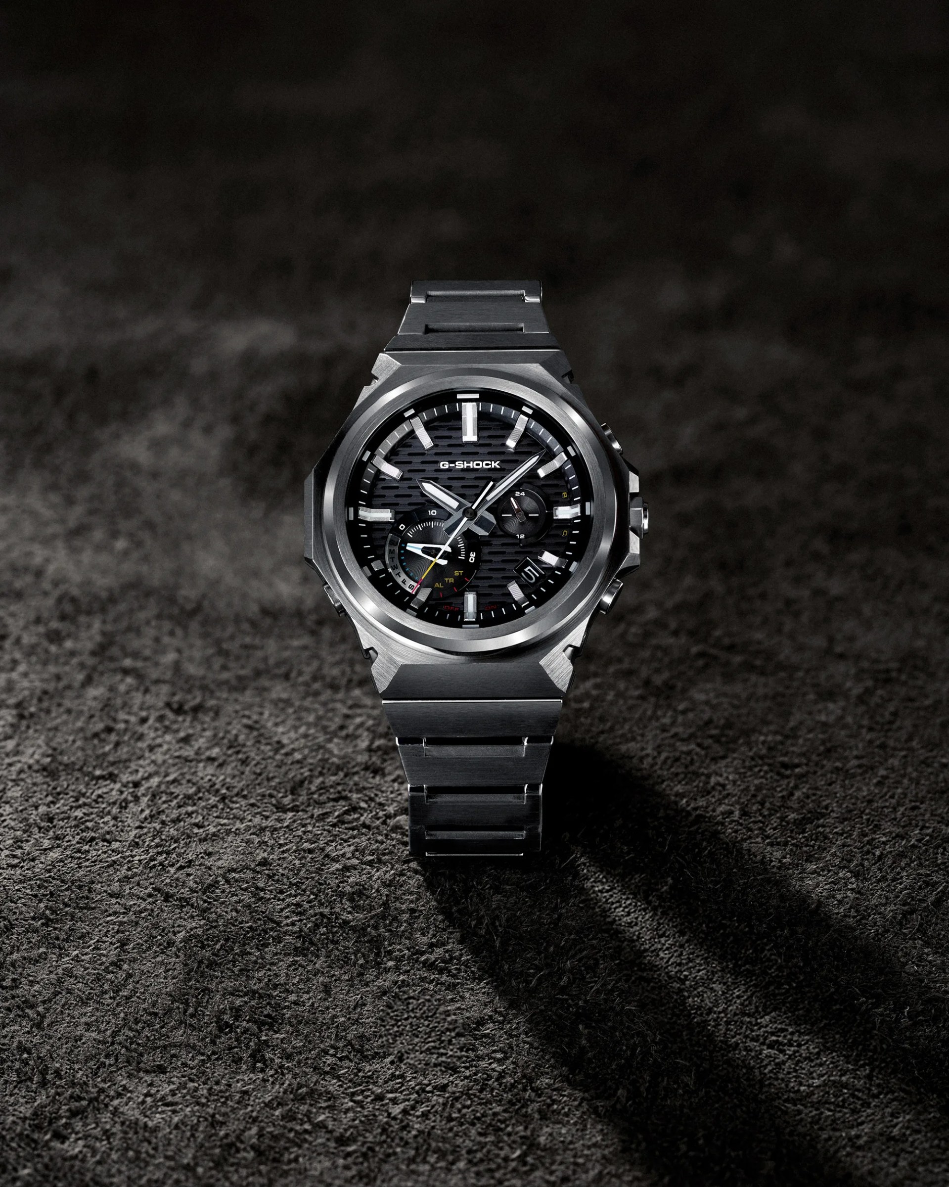 Silver metal G-Shock wristwatch with black textured dial and multiple subdials on a dark textured surface.