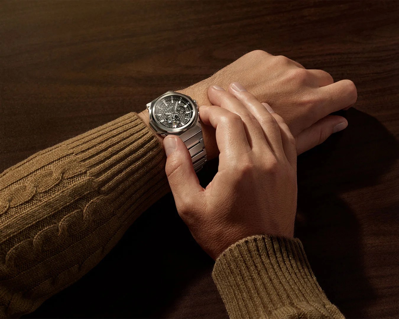 Silver metal wristwatch with a textured dark dial worn on a wrist with a brown cable-knit sweater.