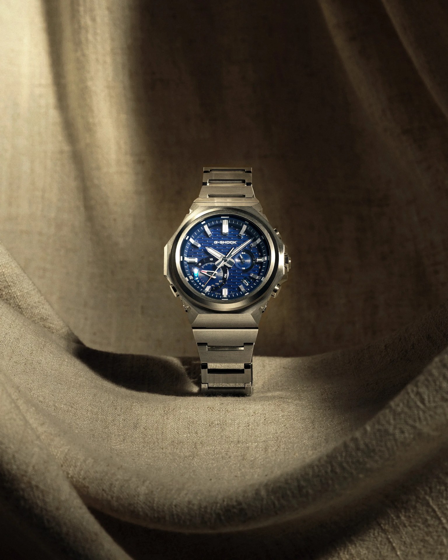 Silver metal G-Shock wristwatch with a blue textured dial on beige fabric background.