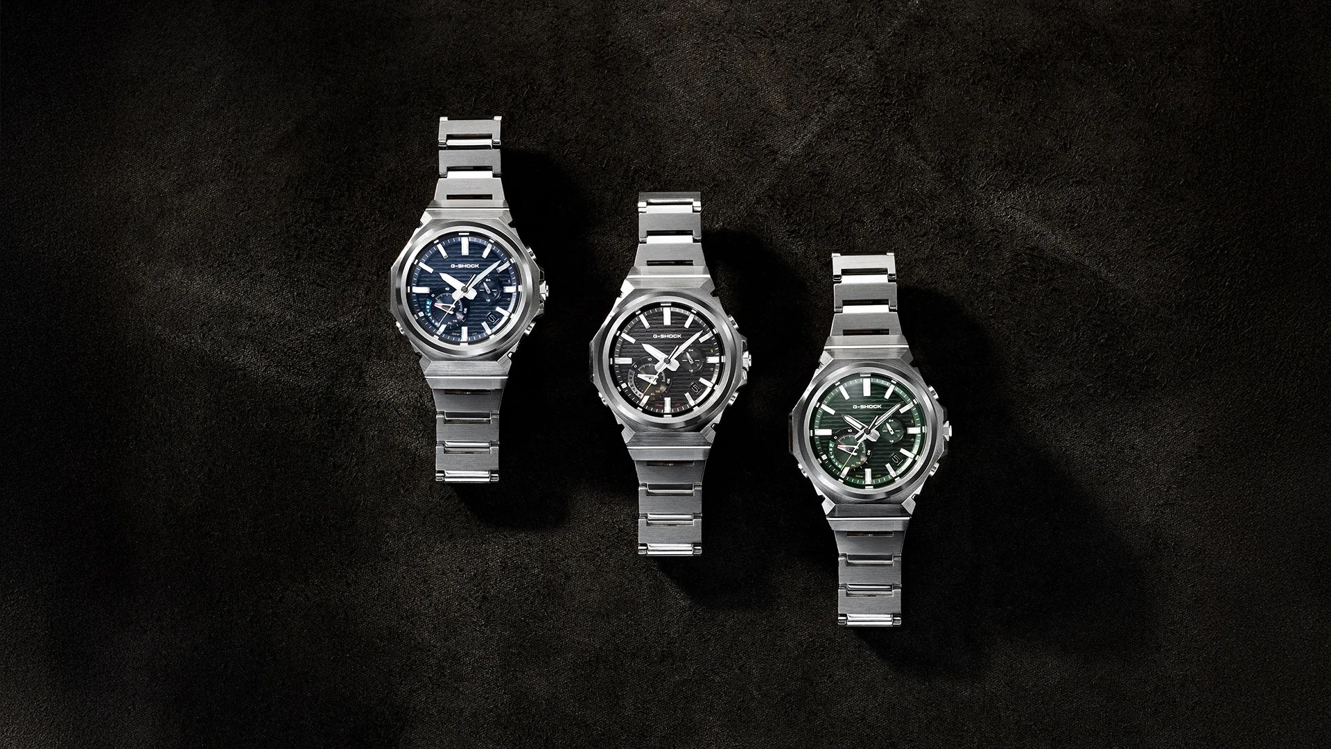 Three stainless steel G-Shock wristwatches with blue, black, and green dials on a dark textured surface.