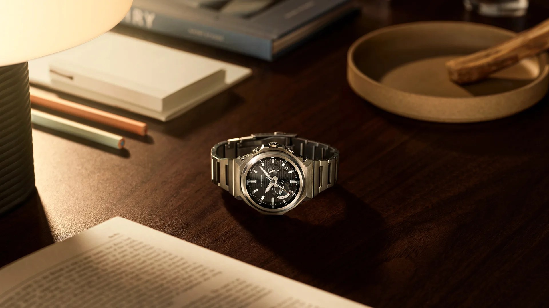 Silver metal G-Shock wristwatch with black dial on a dark wooden desk near a lamp and open book.
