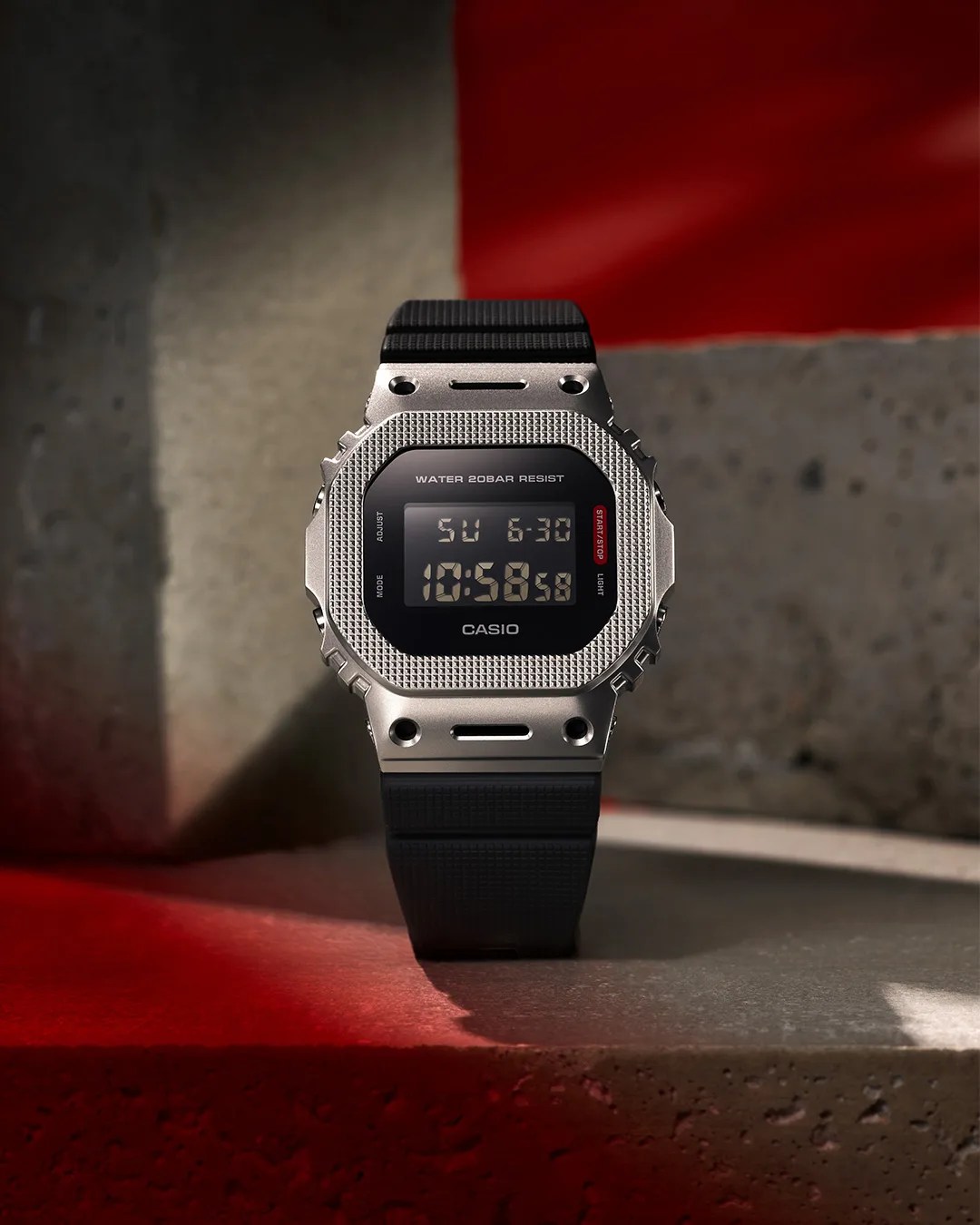 Digital Casio watch with a textured silver bezel and black resin strap on a concrete surface with red lighting.