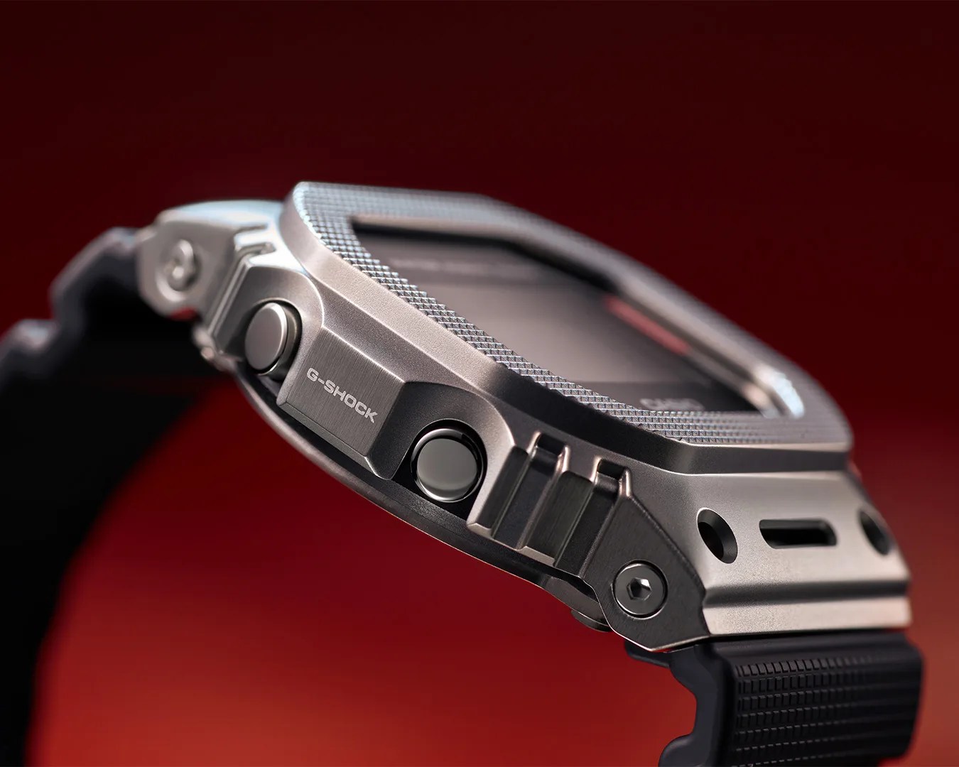 Close-up of a silver metal G-Shock watch case with black buttons and a textured black strap against a red background.