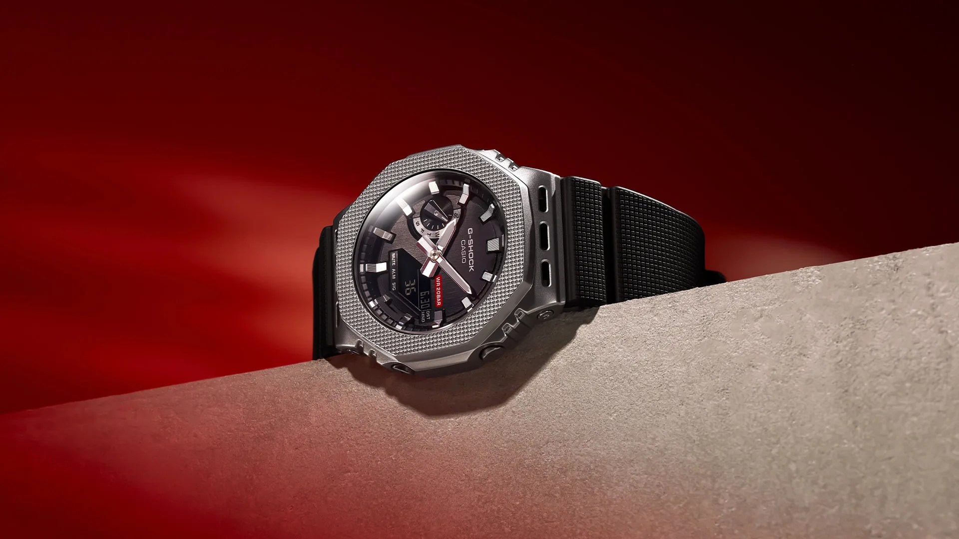 Silver G-Shock Casio watch with black textured strap resting on a beige surface against a red background.