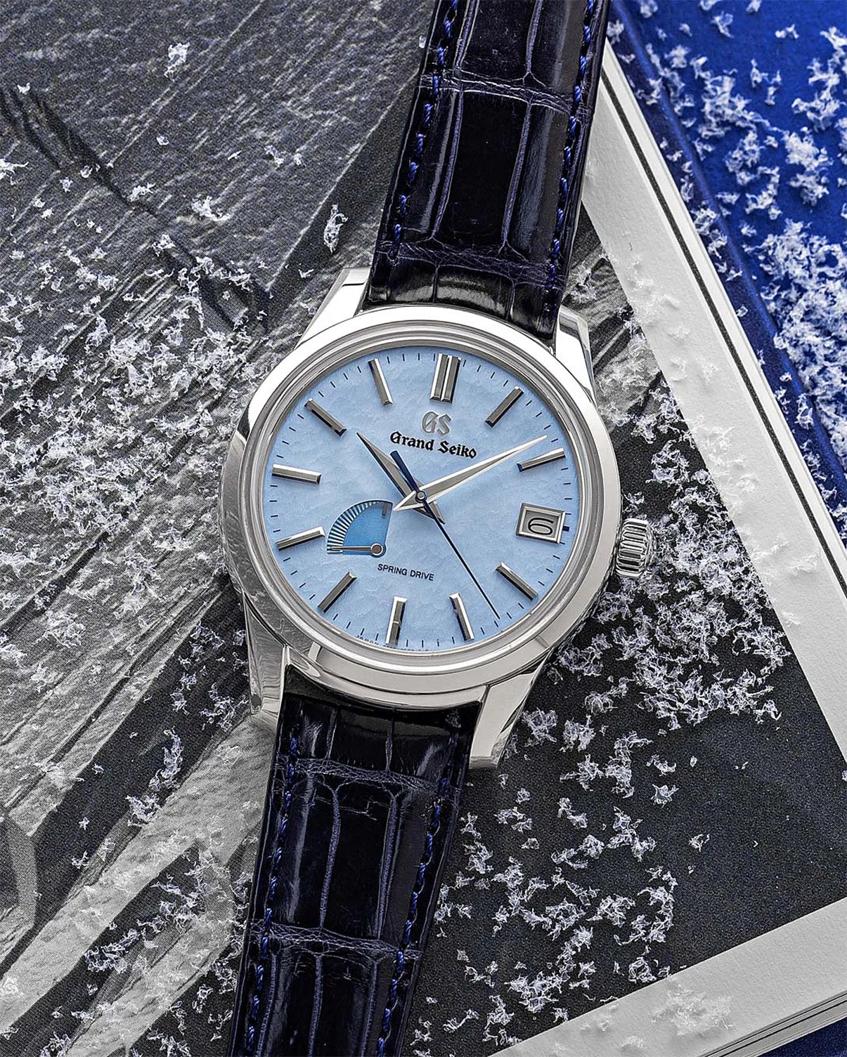 Grand Seiko wristwatch with light blue dial, silver markers, date window, and dark blue leather strap on a snowy surface.