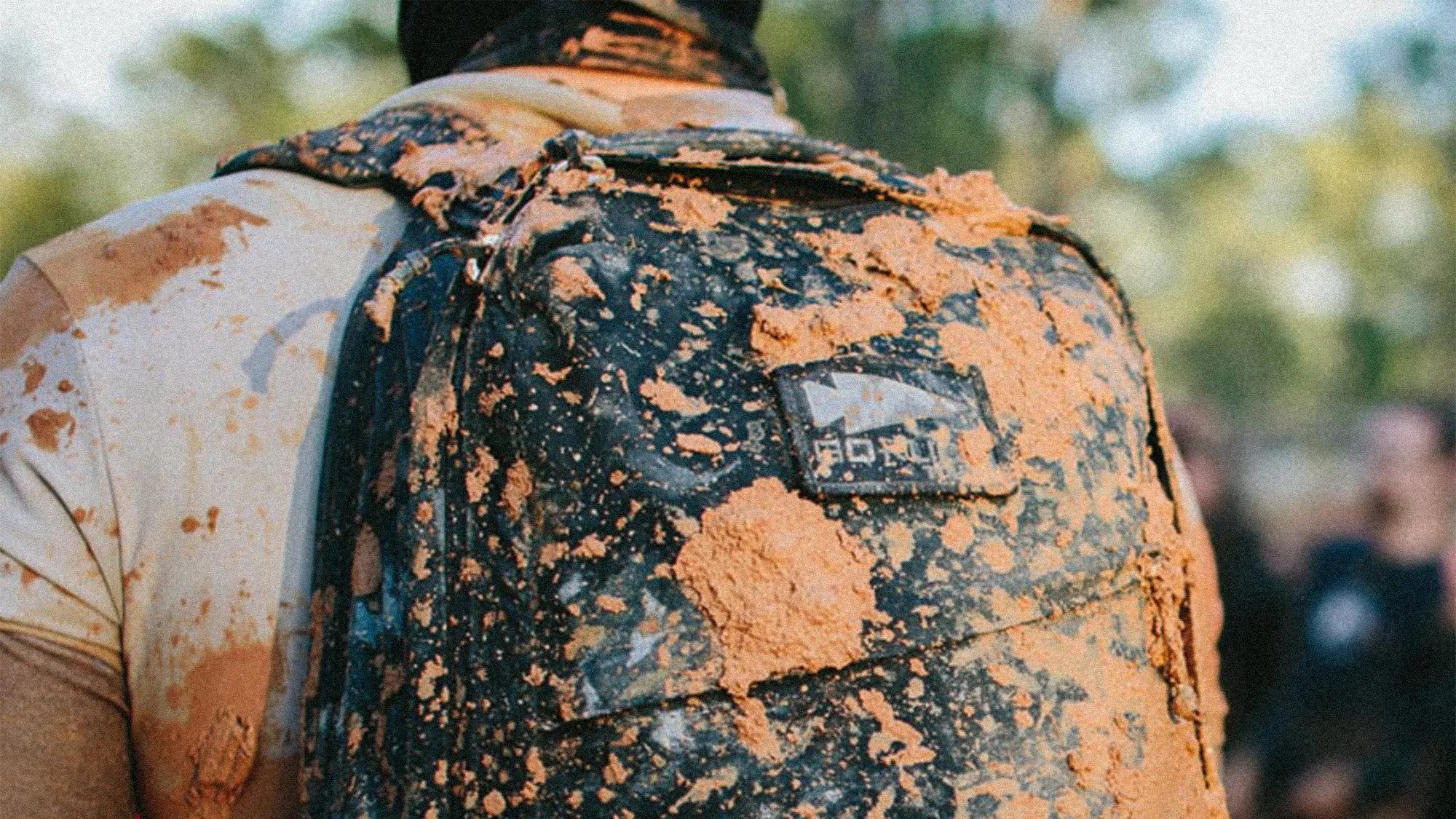 Black backpack covered in orange mud worn by a person in a mud-splattered beige shirt.