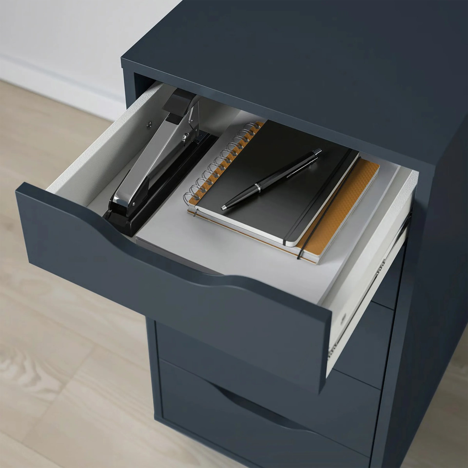Open dark blue drawer with a silver stapler, black pen, and stacked notebooks inside.