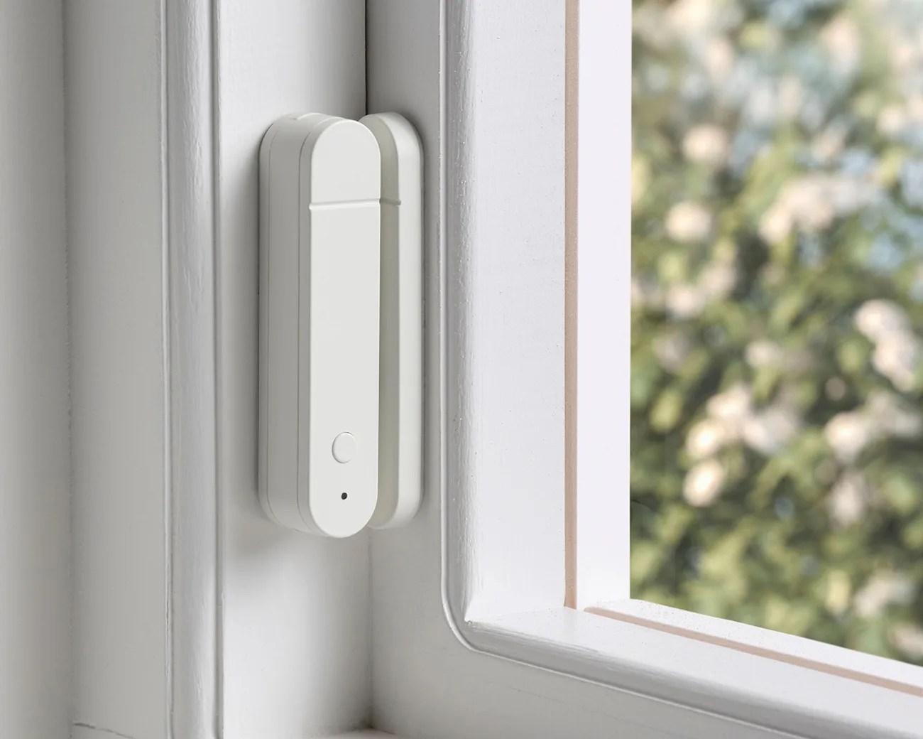 White rectangular window sensor device mounted on a white window frame with blurred greenery outside.