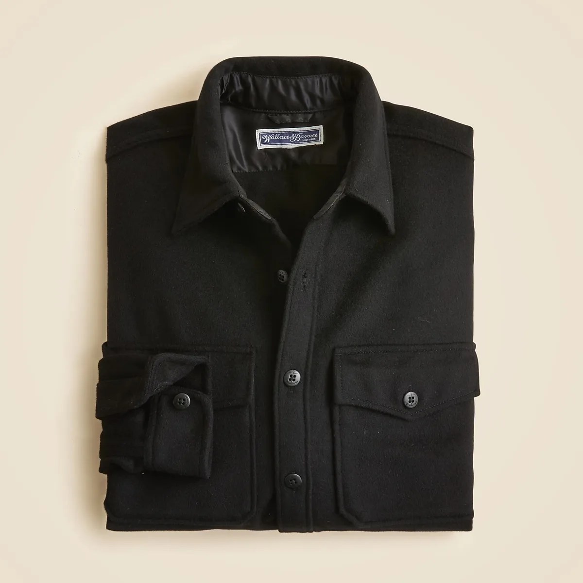 Black button-up wool shirt with two front flap pockets, folded on a beige background.