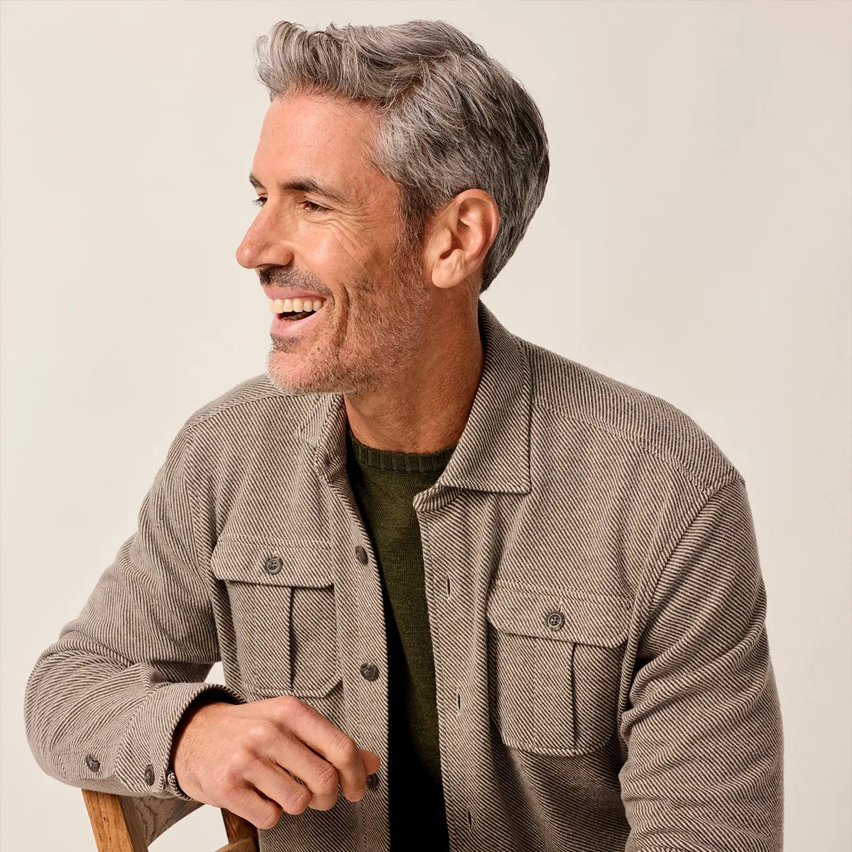 Man with gray hair wearing a beige textured button-up shirt over a dark green sweater, sitting on a wooden chair.