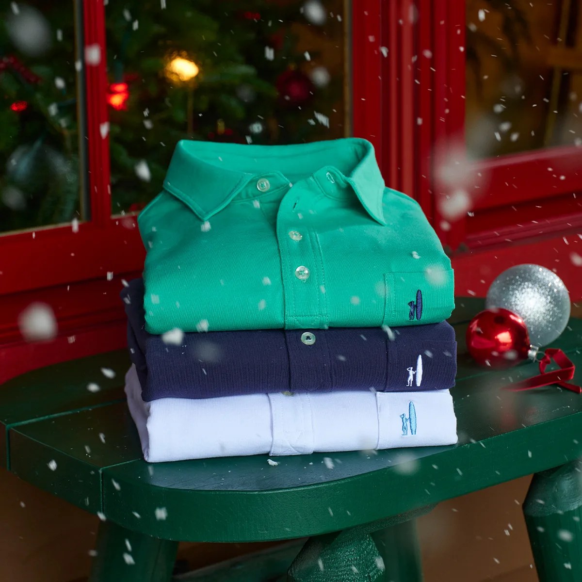Stack of folded green, navy, and white polo shirts on a green wooden table with Christmas ornaments and falling snow.