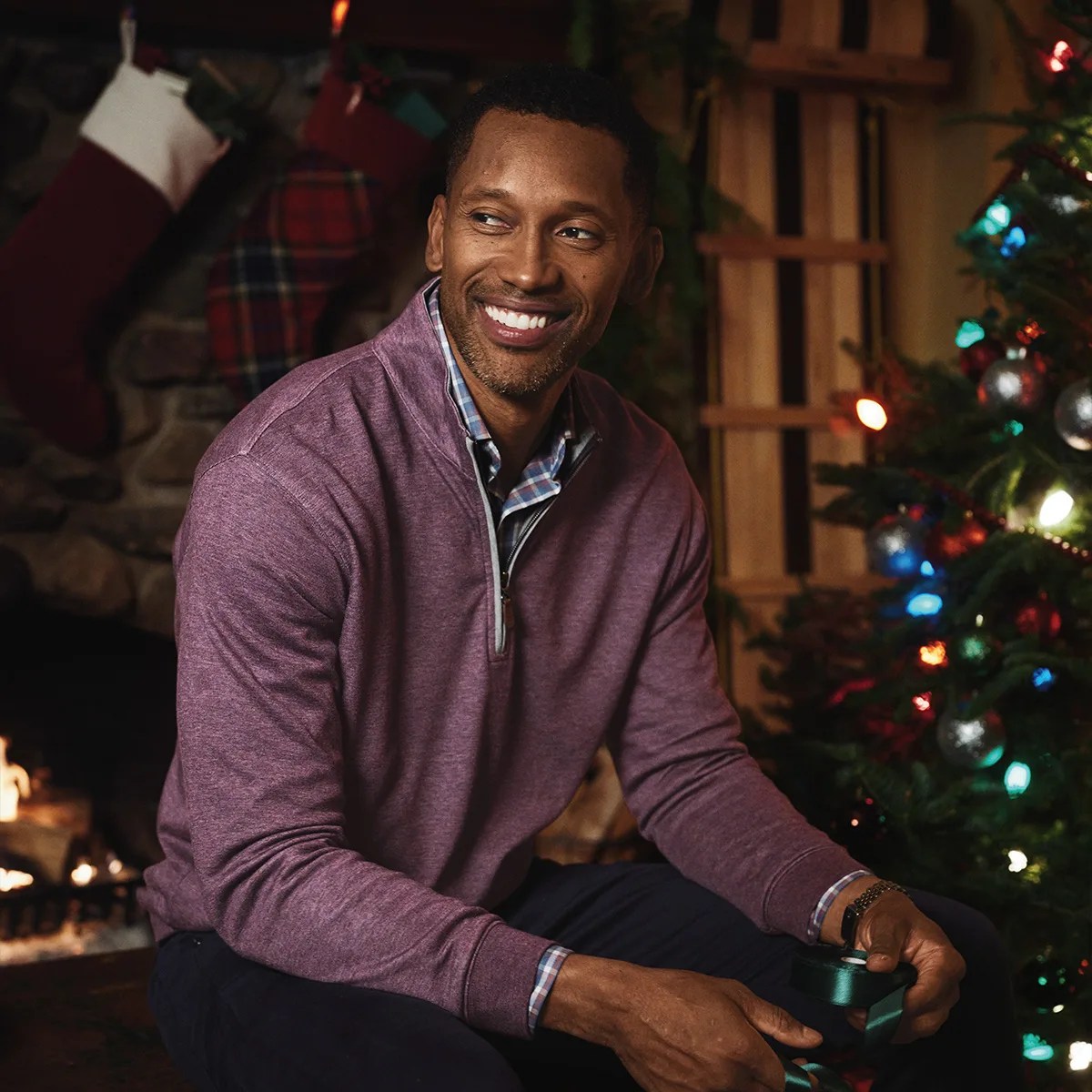 Man wearing a purple quarter-zip sweater over a plaid shirt sitting near a decorated Christmas tree and fireplace.
