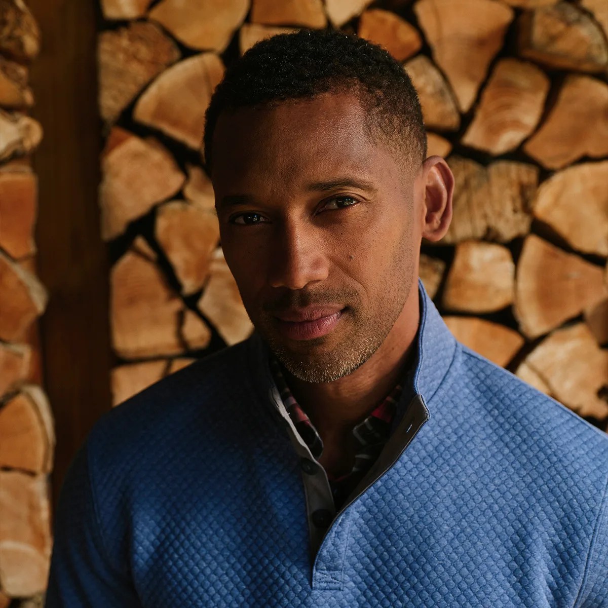 Man wearing a textured blue quarter-zip pullover standing in front of stacked firewood.