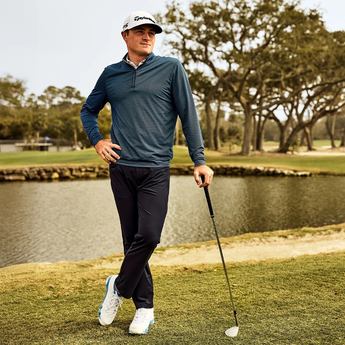 Man wearing a white TaylorMade cap, blue long-sleeve golf shirt, black pants, and white golf shoes standing on a golf course near water holding a golf club.