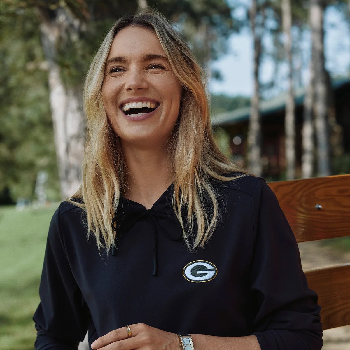 Woman with blonde hair wearing a black hoodie with a Green Bay Packers logo sitting on a wooden bench outdoors.