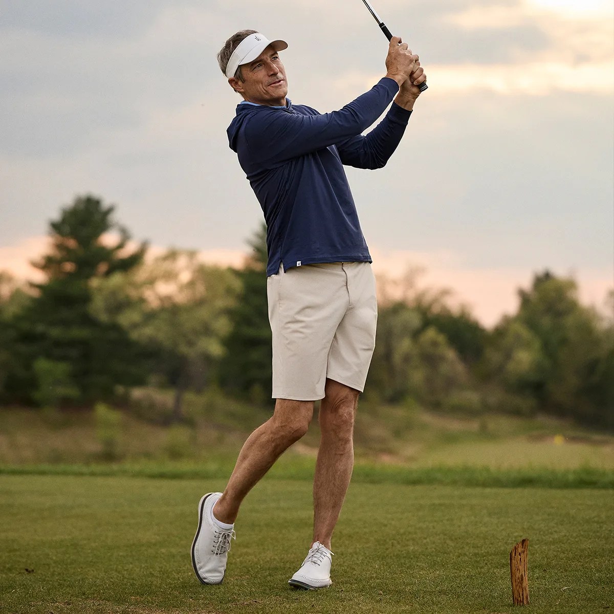 Man wearing navy long-sleeve shirt, beige shorts, white golf shoes, and white visor swinging golf club on a course.