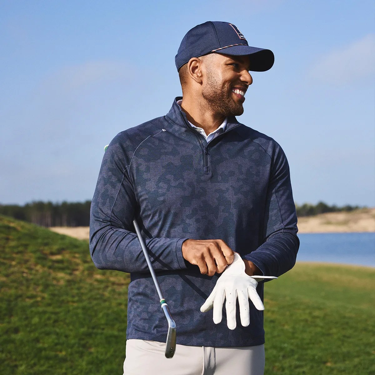 Man wearing a navy patterned quarter-zip pullover and navy cap putting on a white golf glove on a golf course.