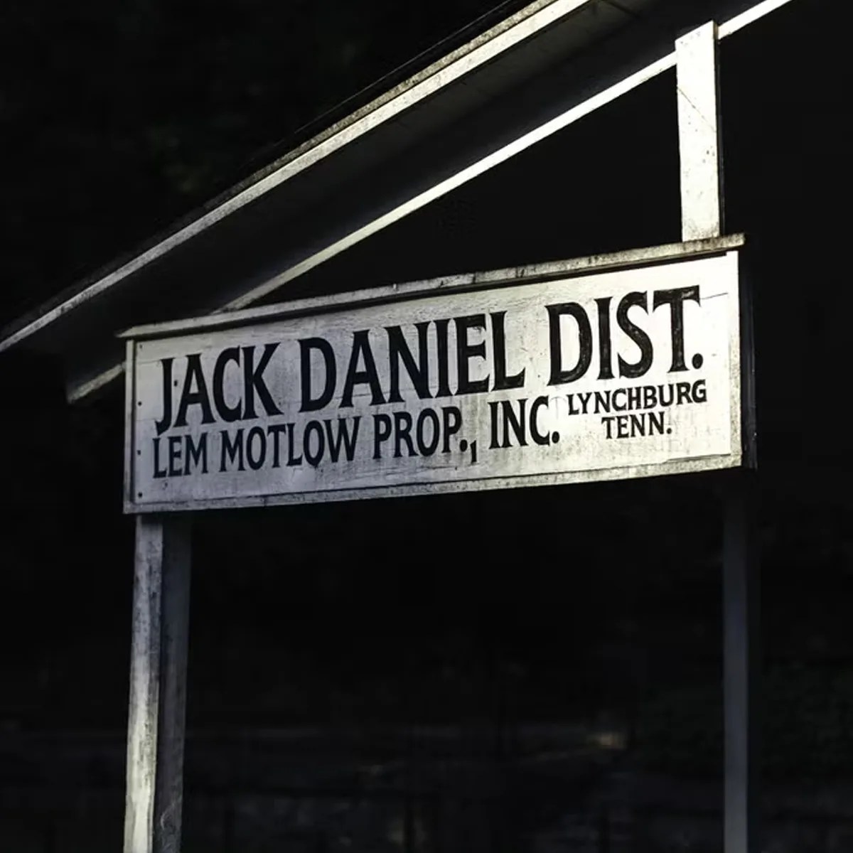 White wooden sign with black text reading "JACK DANIEL DIST. LEM MOTLOW PROP., INC. LYNCHBURG TENN." against a dark background.