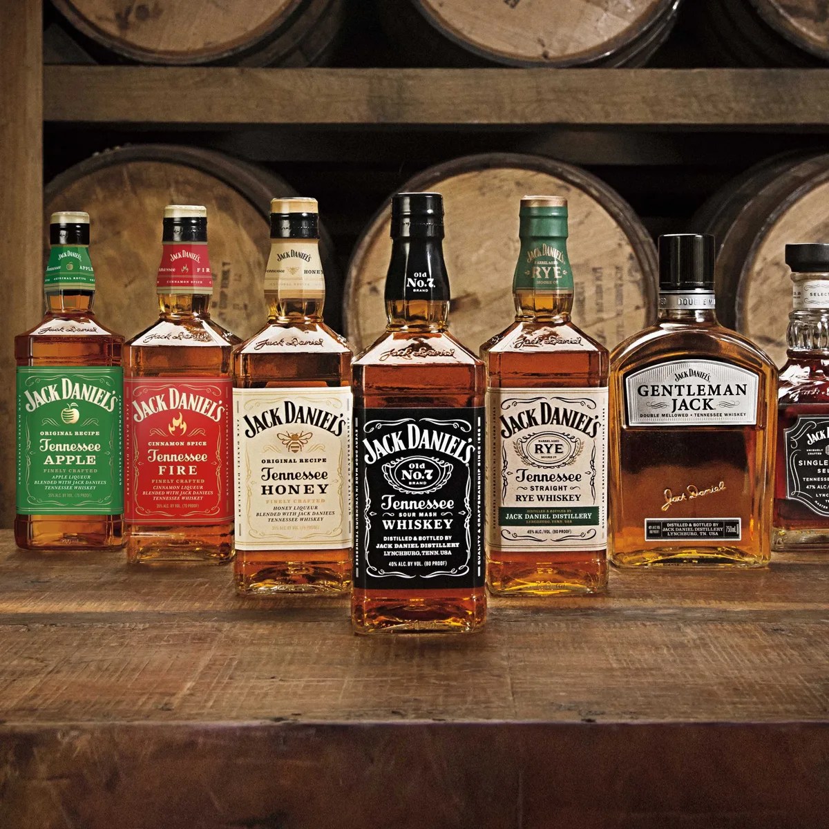 Seven bottles of Jack Daniel's whiskey varieties including Tennessee Apple, Tennessee Fire, Tennessee Honey, Old No. 7, Tennessee Rye, Gentleman Jack, and Single Barrel Select on a wooden surface with barrels in the background.