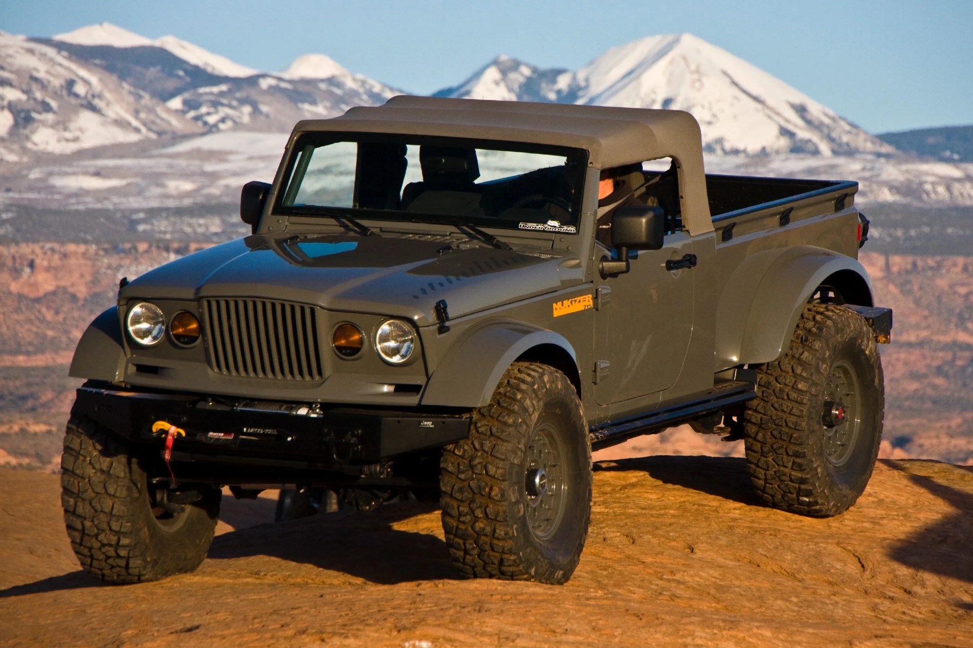 Gray off-road pickup truck with large tires parked on rocky terrain with snow-capped mountains in the background.