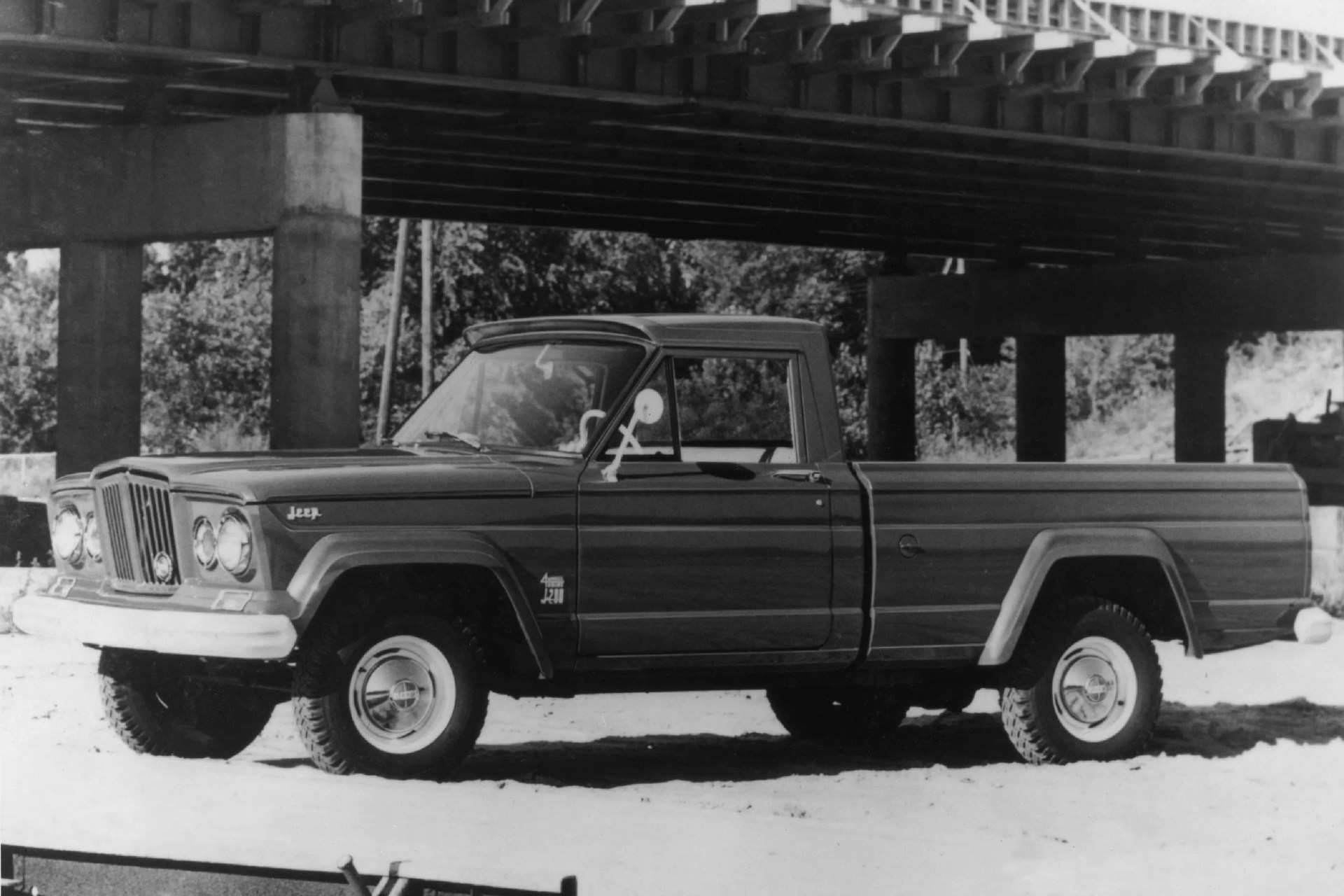 Black and white photo of a classic Jeep J-10 pickup truck parked under a bridge.