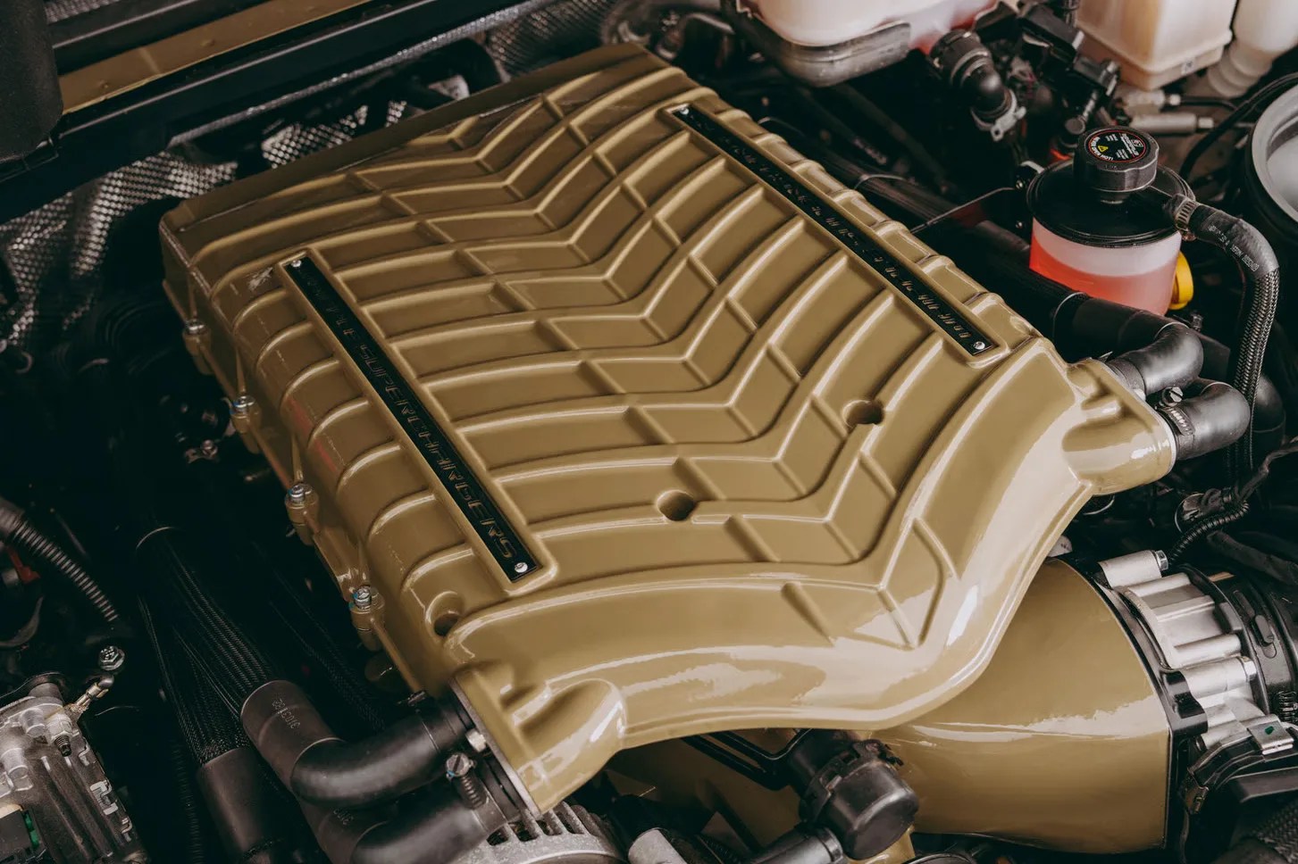 Glossy beige supercharger unit with black hoses and metal components in a car engine bay.