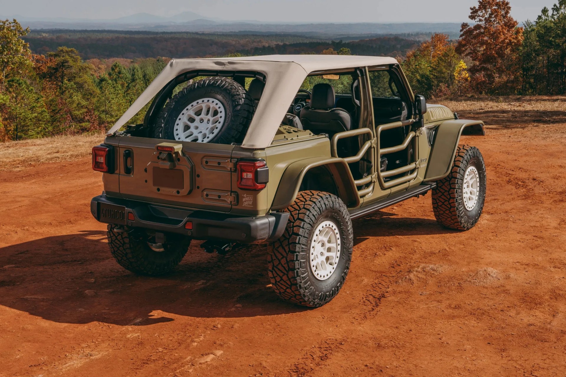 Olive green Jeep with beige soft top and tubular doors parked on a dirt trail with forest in the background.