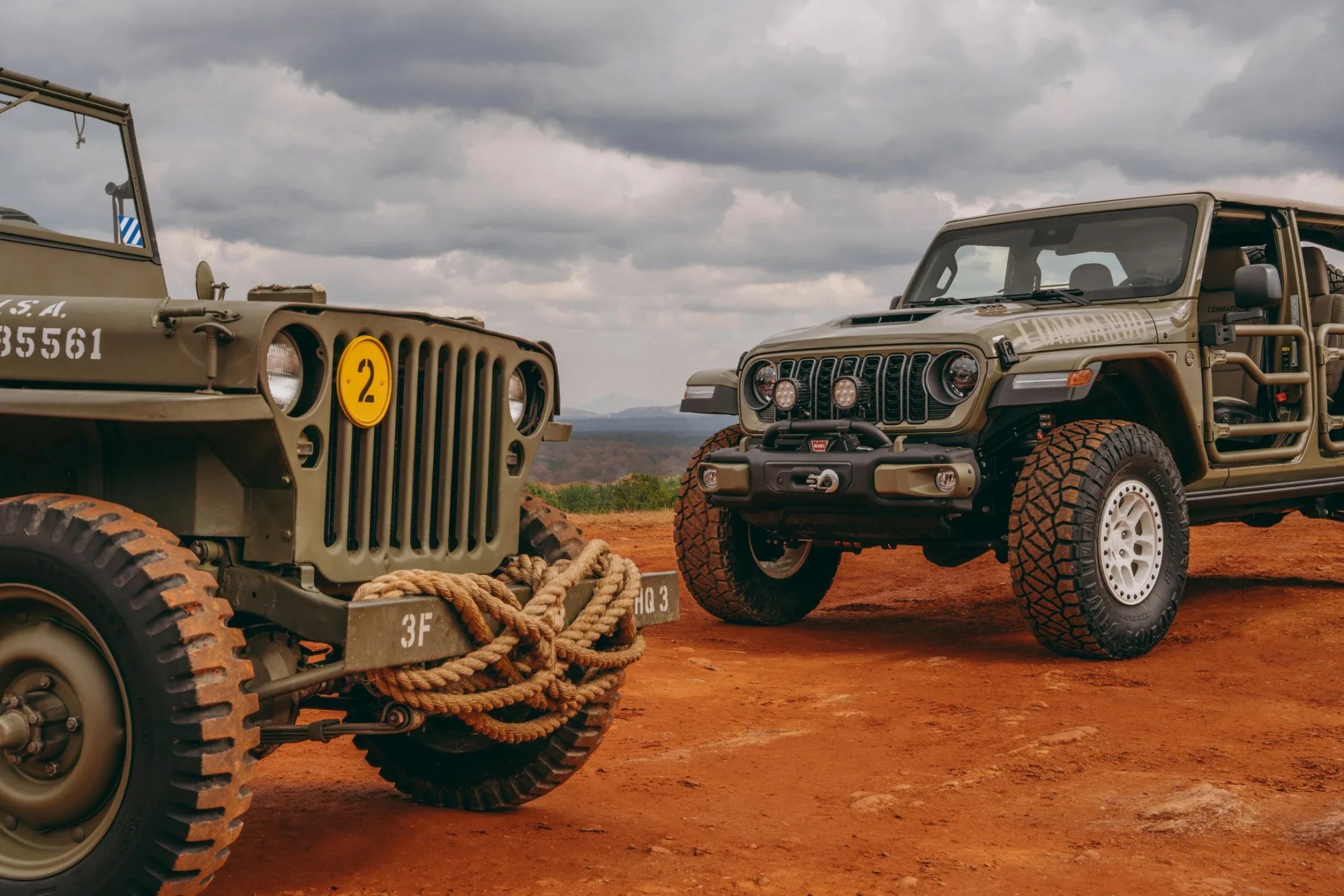 Two olive-green off-road vehicles, one vintage military jeep with a rope on the front bumper and one modern open-door Jeep Wrangler, parked on red dirt under a cloudy sky.