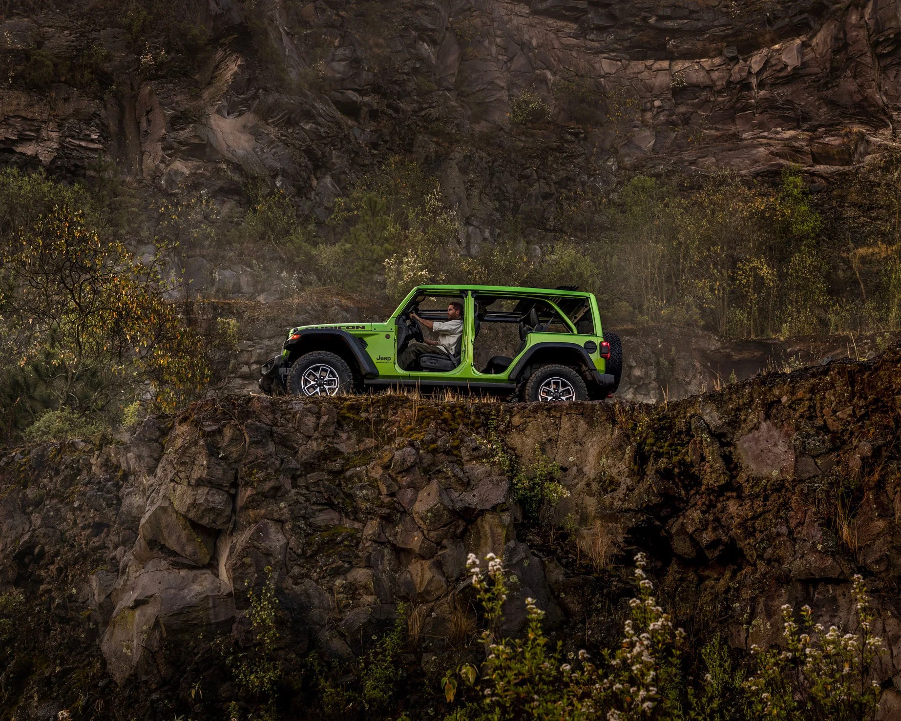 Bright green Jeep Rubicon with open doors driven on rocky cliffside with dense foliage and rugged rock formations.