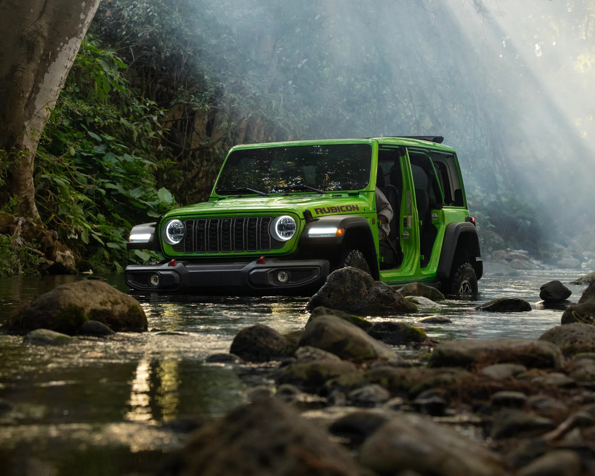 Bright green Jeep Rubicon with open doors driving through a shallow rocky stream in a forest.