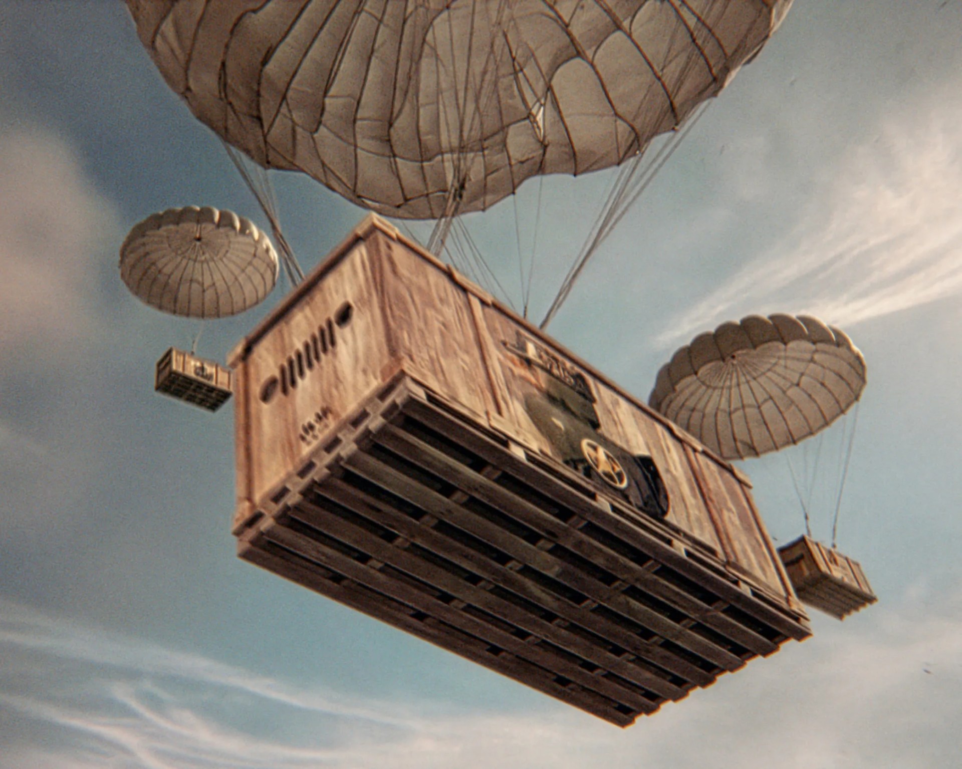 Large wooden crate and two smaller crates suspended by parachutes against a blue sky with clouds.