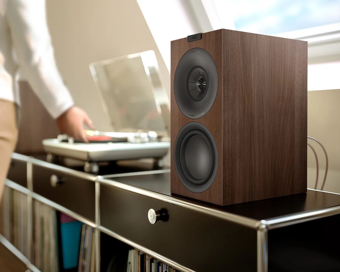 Wood-finished KEF speaker with black drivers on a black metal shelf near a person using a turntable.