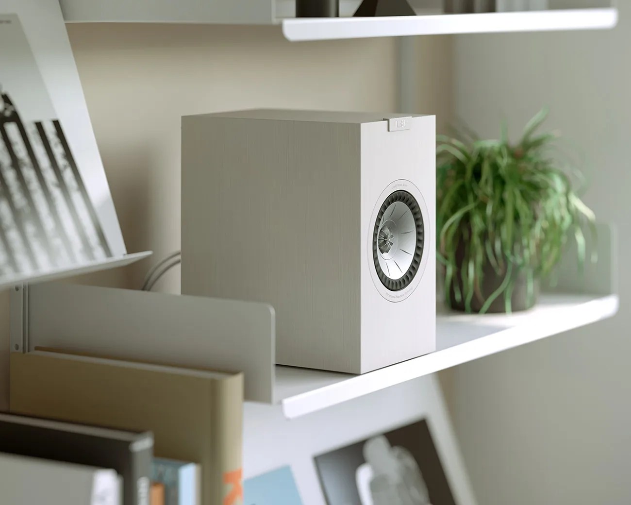 White KEF speaker with silver driver on a white shelf next to a green potted plant and books.