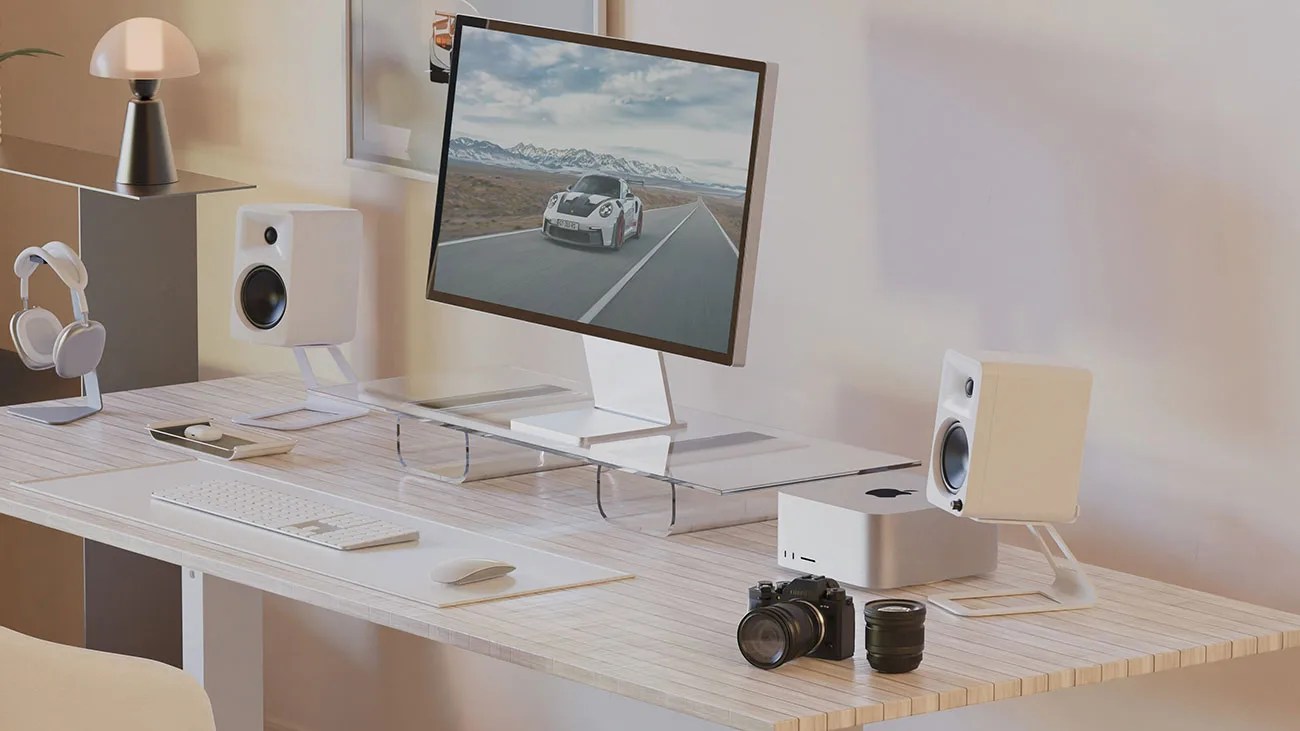 Minimalist desk setup with a monitor on a clear stand, white speakers, keyboard, mouse, camera, and headphones.