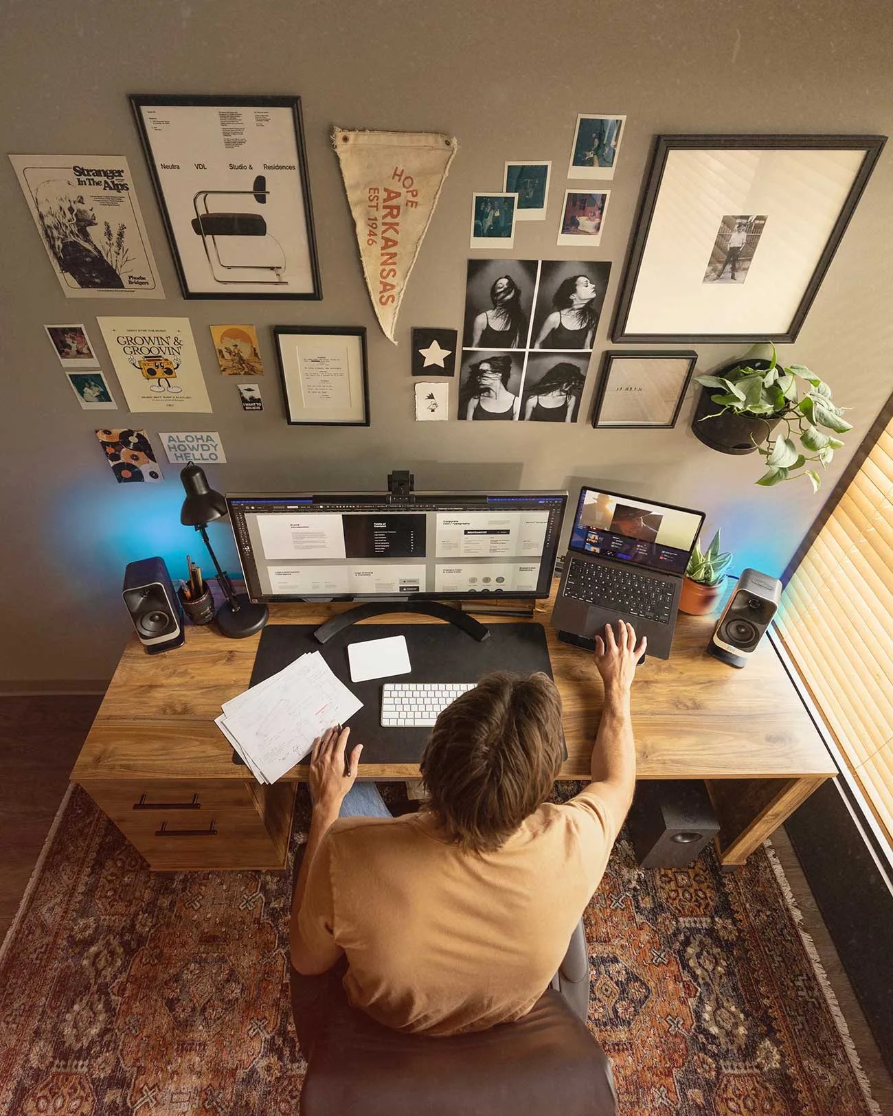 Overhead view of a person working at a wooden desk with an ultrawide monitor, laptop, papers, and wall art above.