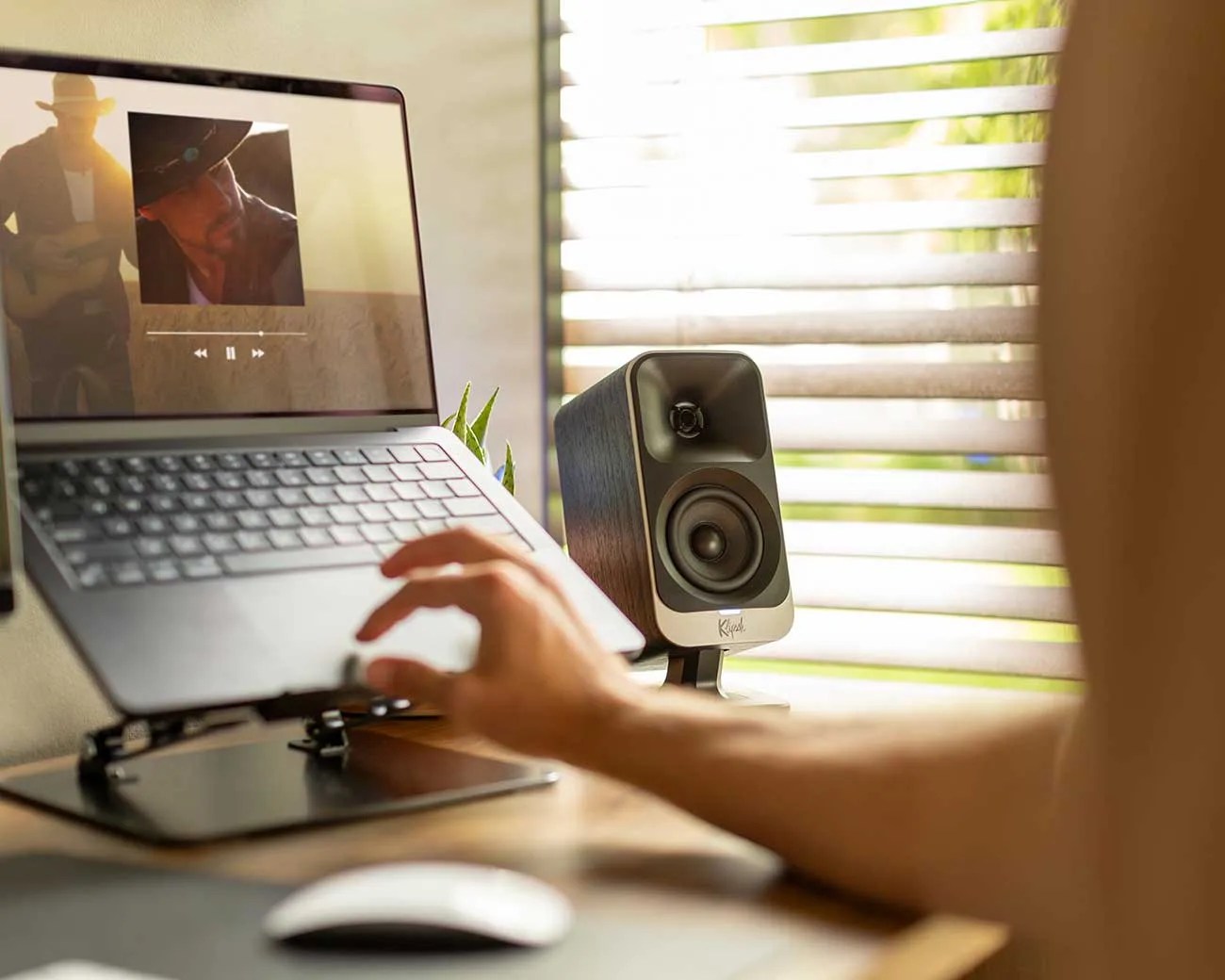 Black Klipsch speaker on a desk next to a laptop with a music player on screen and a hand reaching toward the keyboard.