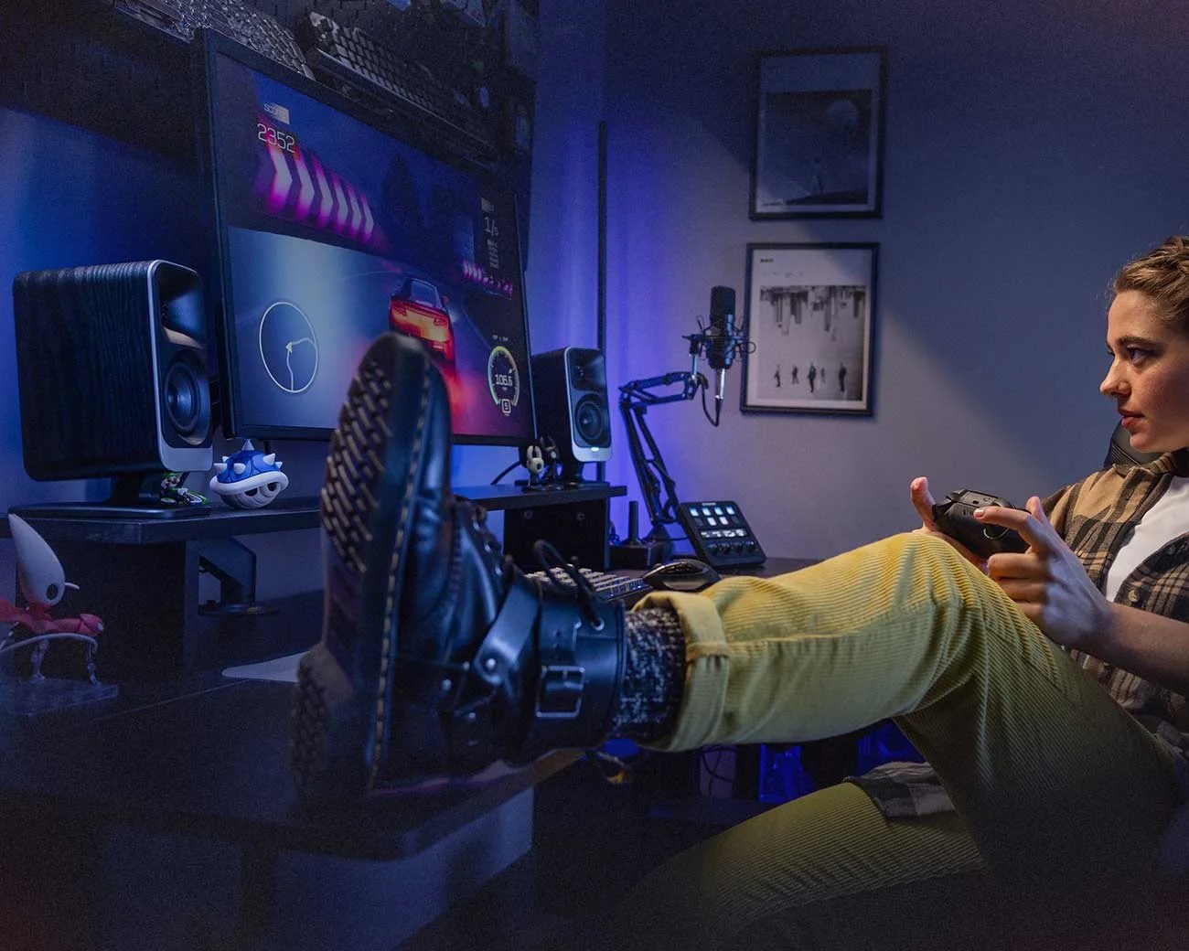Person wearing yellow pants and black boots playing a racing video game on a monitor with speakers and a microphone on the desk.