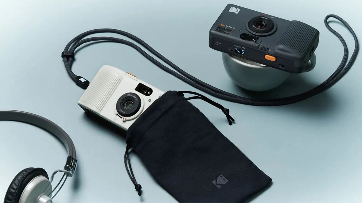 Two Kodak compact cameras, one black resting on a small bowl and one white partially inside a black drawstring pouch, both with neck straps.