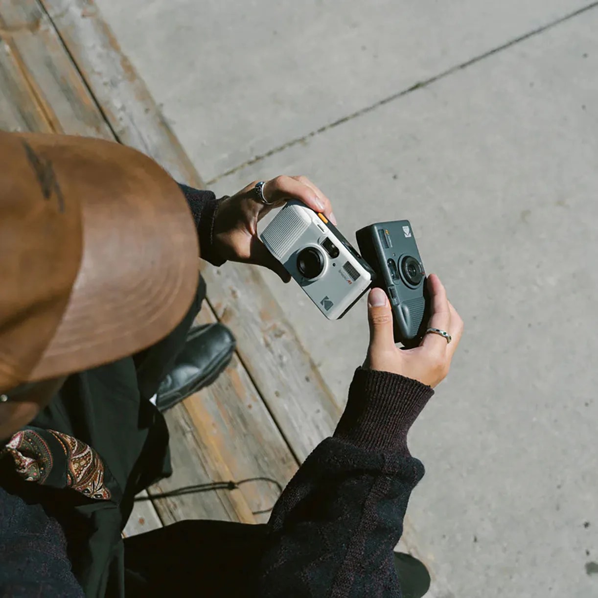 Person wearing a brown cap and dark sweater holding two compact Kodak cameras, one white and one black.