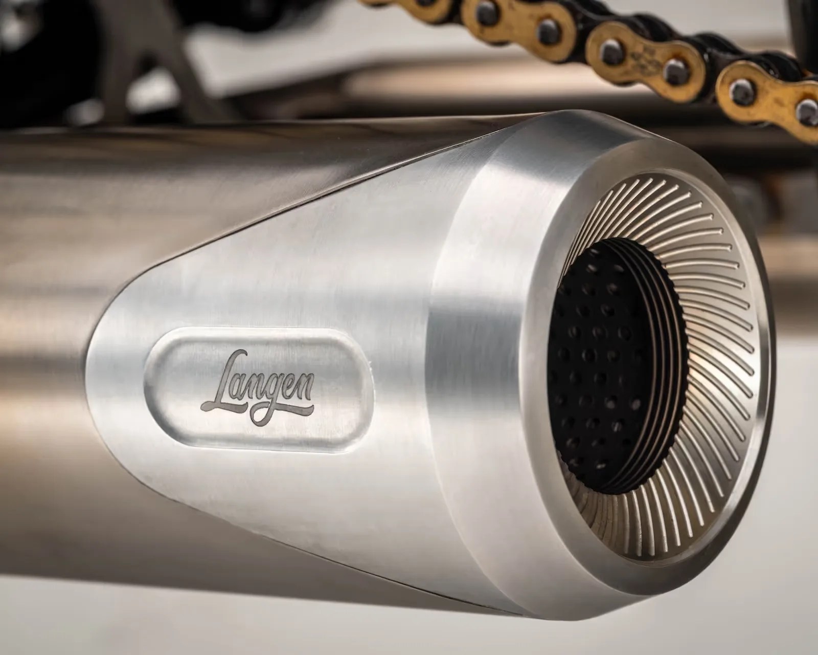 Brushed stainless steel cylindrical exhaust pipe with "Langen" engraved and a gold bike chain above it.