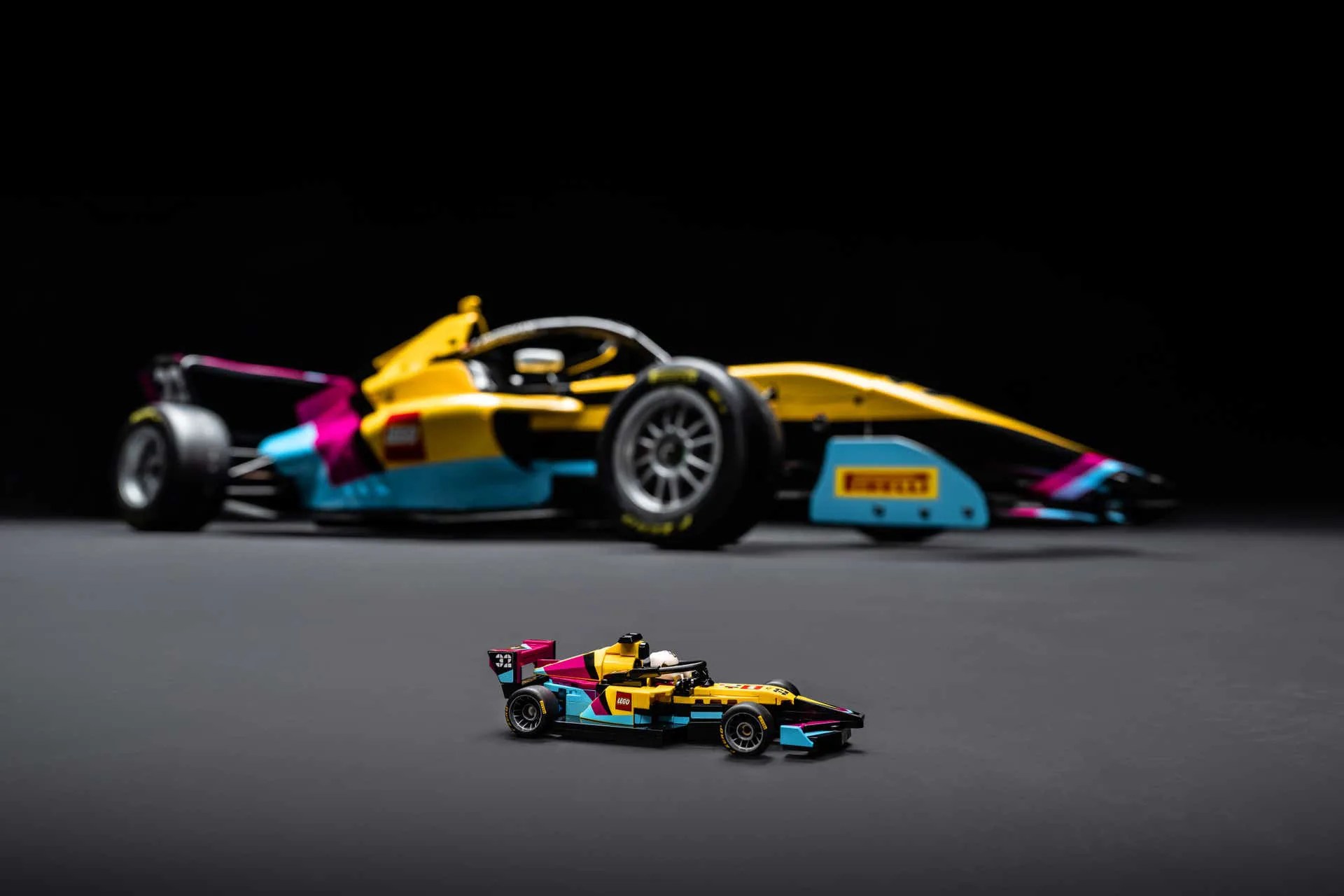 Yellow, blue, and pink LEGO Formula 1 car model in front of a full-sized matching race car on a gray surface.