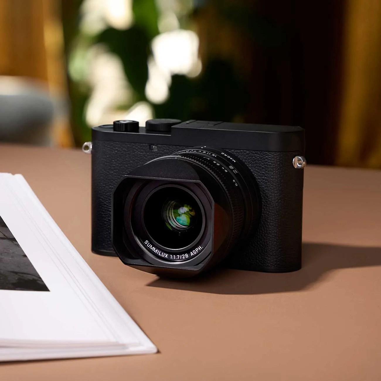 Black compact camera with textured body and a Summilux 28mm f/1.7 lens on a brown surface next to a book.