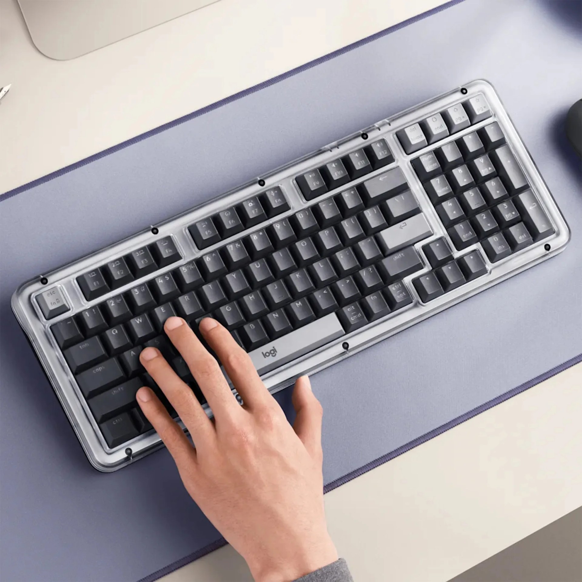 Hand resting on a black and gray Logitech mechanical keyboard with a clear protective cover on a blue desk mat.
