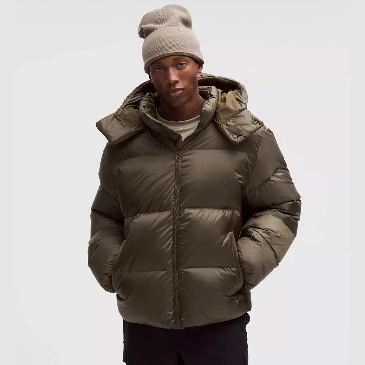 Man wearing an olive green puffer jacket with a hood and a beige knit beanie.
