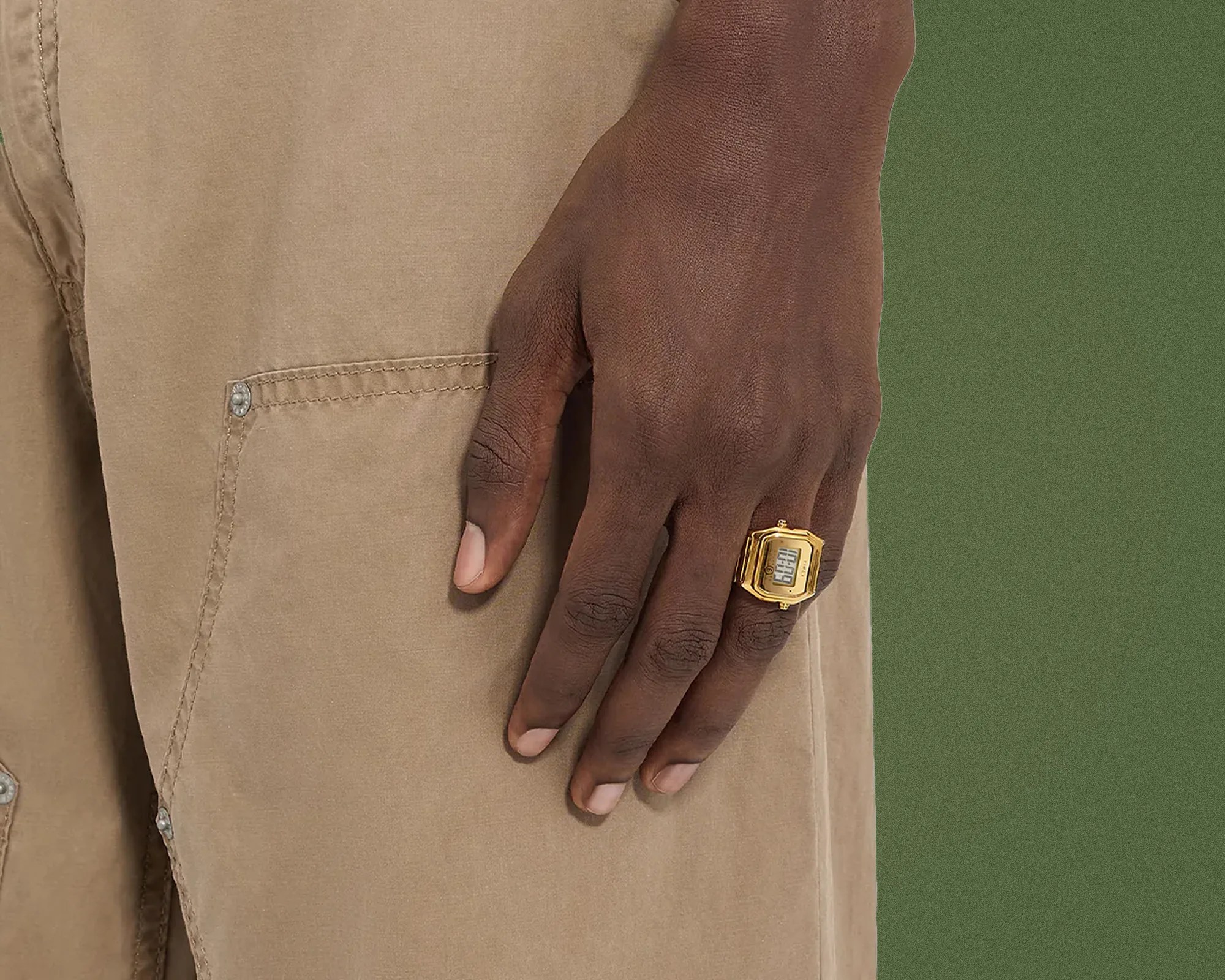 Hand wearing a gold digital watch ring resting on beige pants with visible stitching and metal rivets.