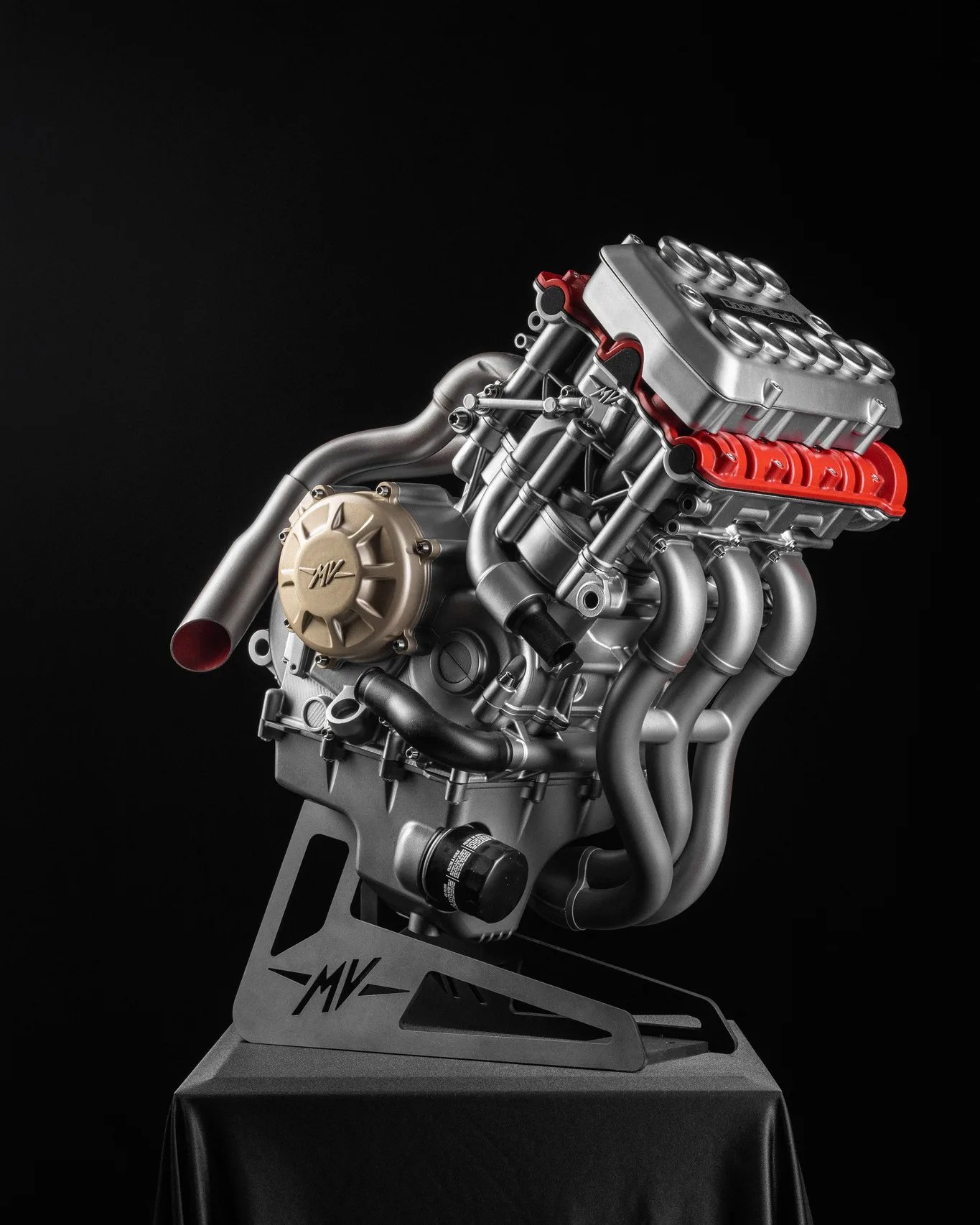 Silver and red MV Agusta motorcycle engine mounted on a black stand with MV logo against a black background.