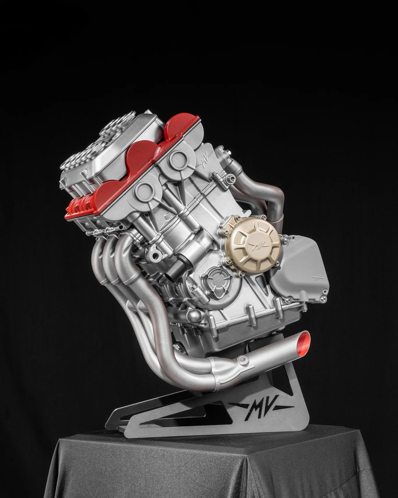 Silver motorcycle engine with red and gold accents mounted on a black stand with "MV" logo against a black background.