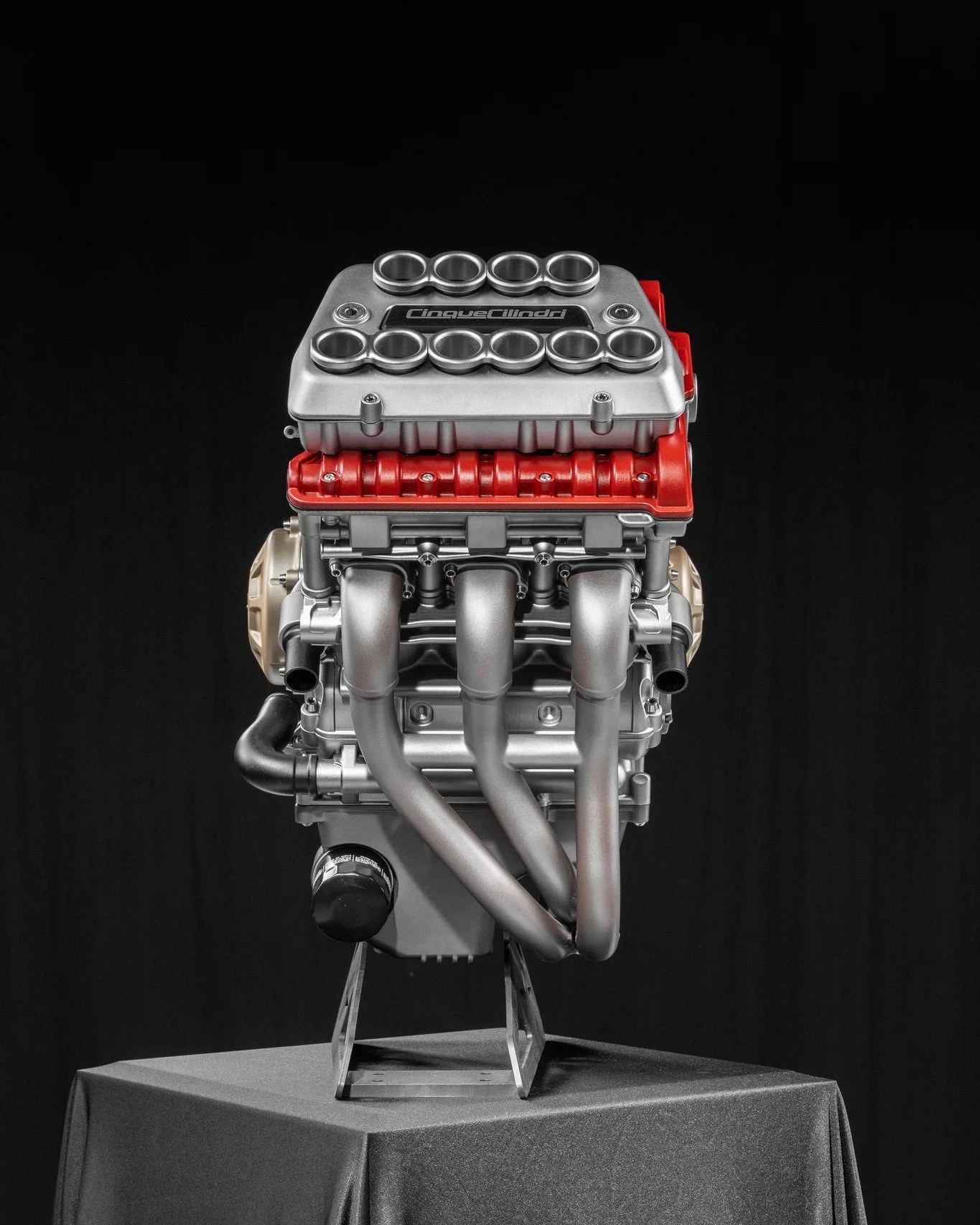 Silver and red Cinque Cilindri engine displayed on a black cloth-covered pedestal against a black background.