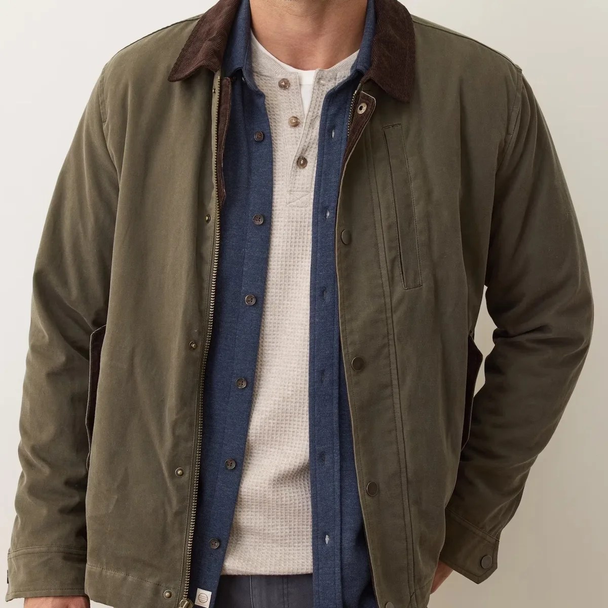 Olive green jacket with brown corduroy collar worn over a blue button-up shirt and cream waffle-knit henley.